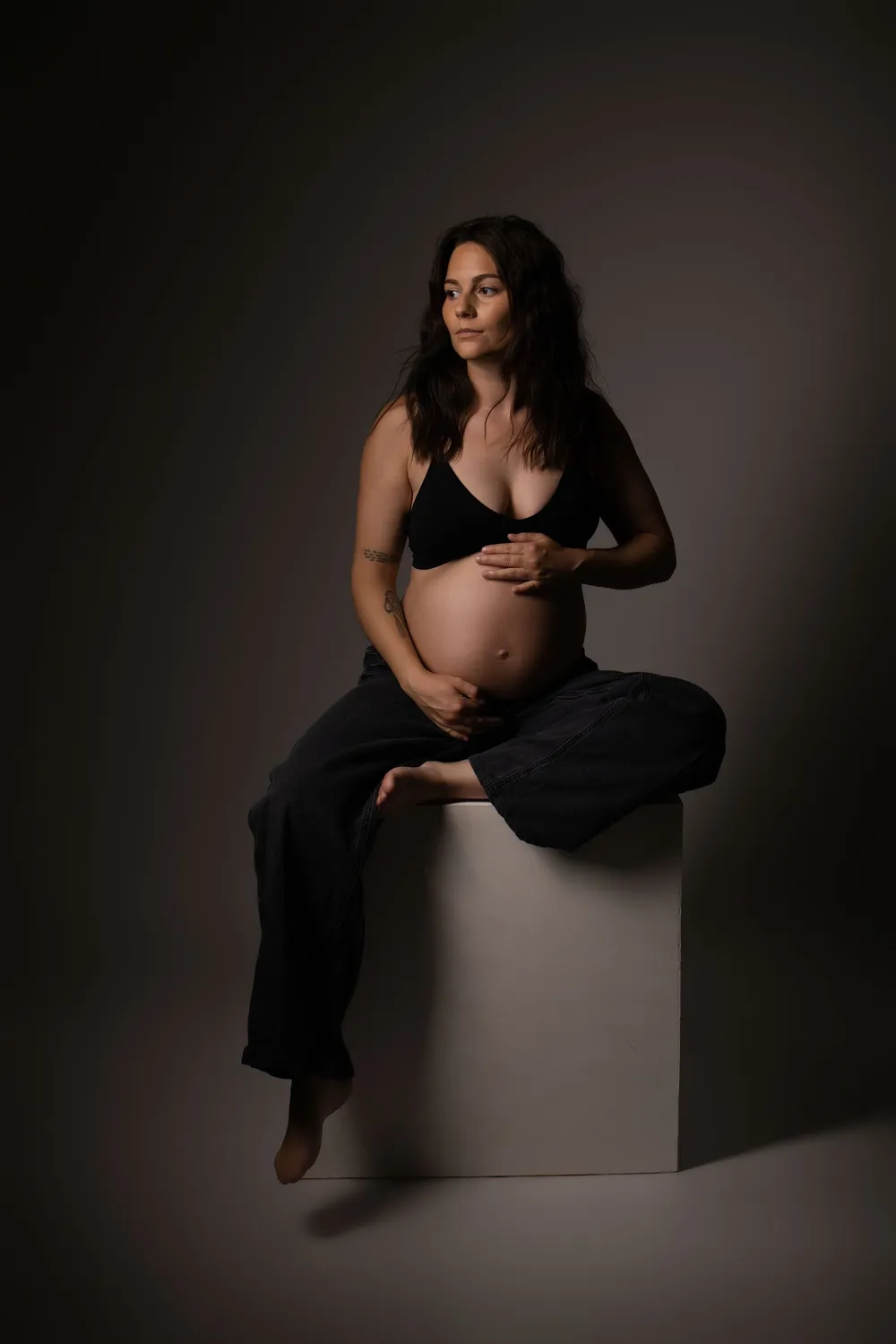 Femme enceinte assise sur un cube blanc, mains sur son ventre rond, séance grossesse en studio - Cindy Poulet photographe