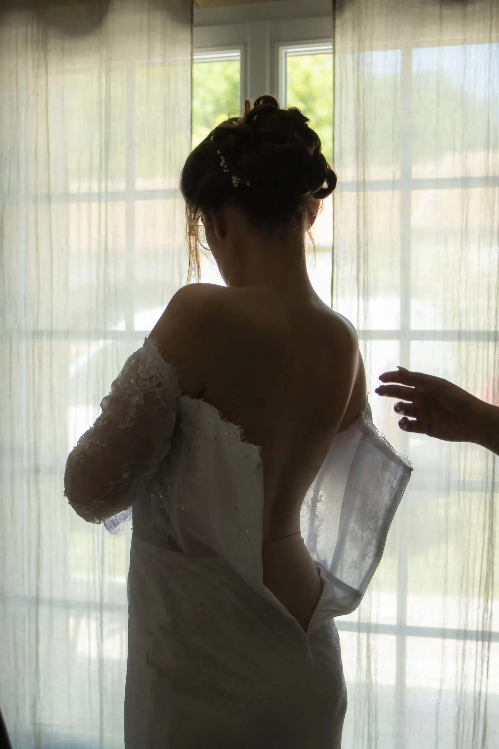 Mariée de dos que l'on lace dans sa robe devant une fenêtre, préparatifs de mariage - Cindy Poulet photographe