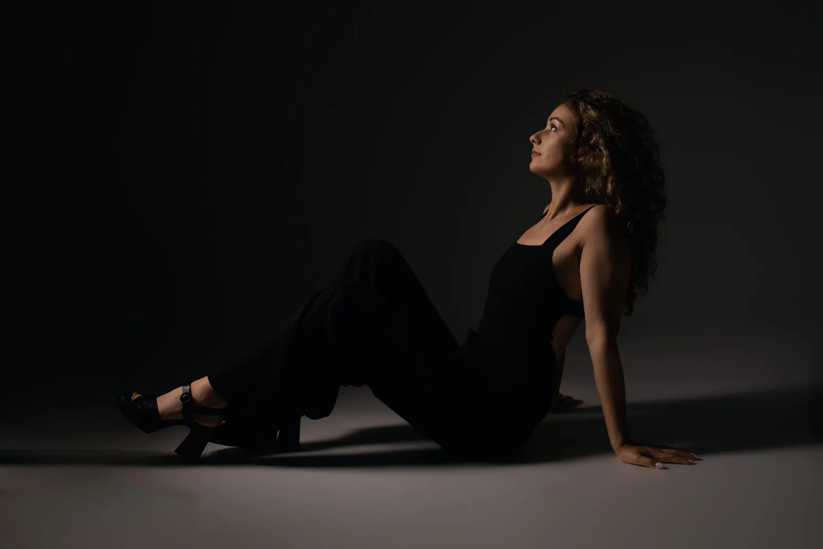 Femme en tenue noire assise au sol, regard levé dans la pénombre du studio - Cindy Poulet photographe