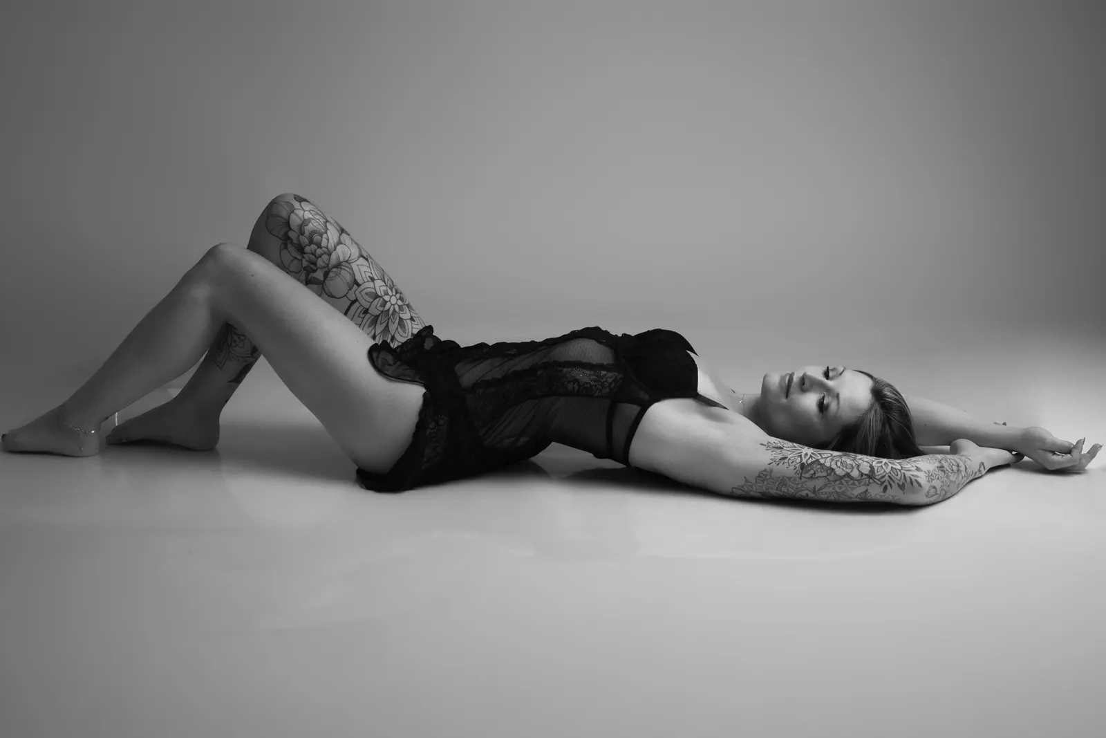 Femme tatouée en body de dentelle noire allongée au sol, bras au-dessus de la tête, boudoir en noir et blanc - Cindy Poulet photographe