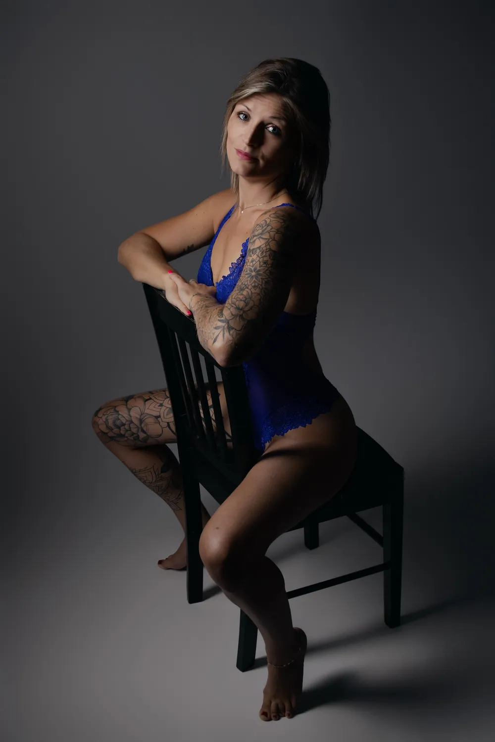 Femme tatouée en body de dentelle bleue assise à califourchon sur une chaise noire, portrait boudoir en studio - Cindy Poulet photographe
