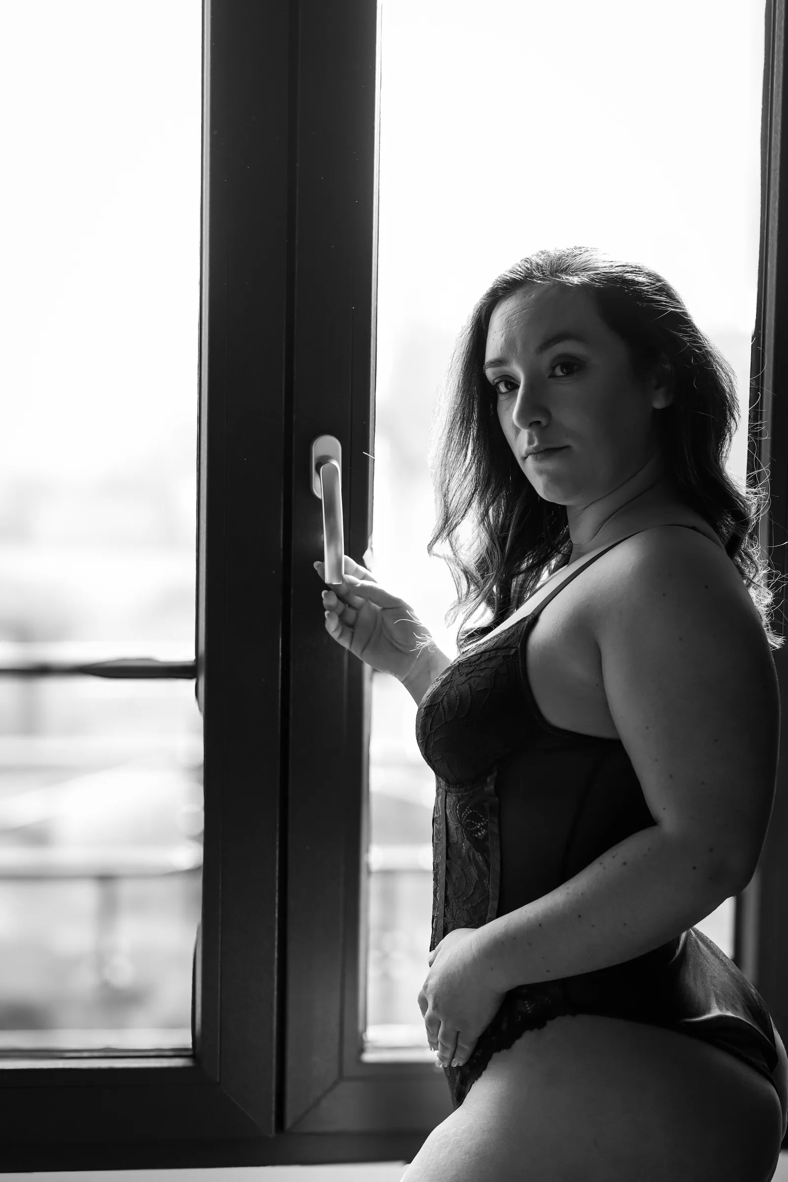Femme en body de dentelle noire près d'une fenêtre, regard vers l'objectif, portrait boudoir en noir et blanc - Cindy Poulet photographe