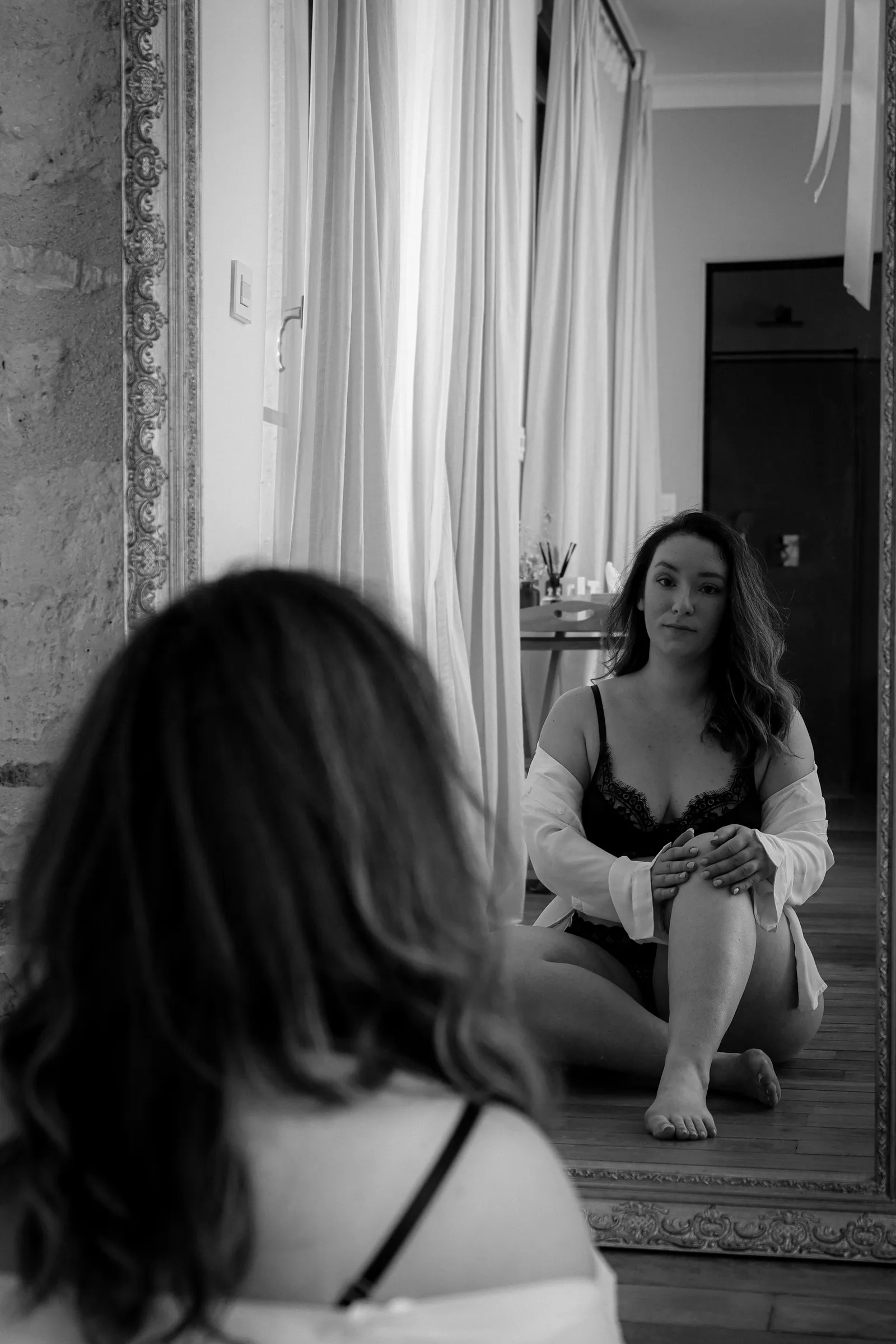 Femme en lingerie noire assise au sol face à un grand miroir ancien, séance boudoir en noir et blanc - Cindy Poulet photographe