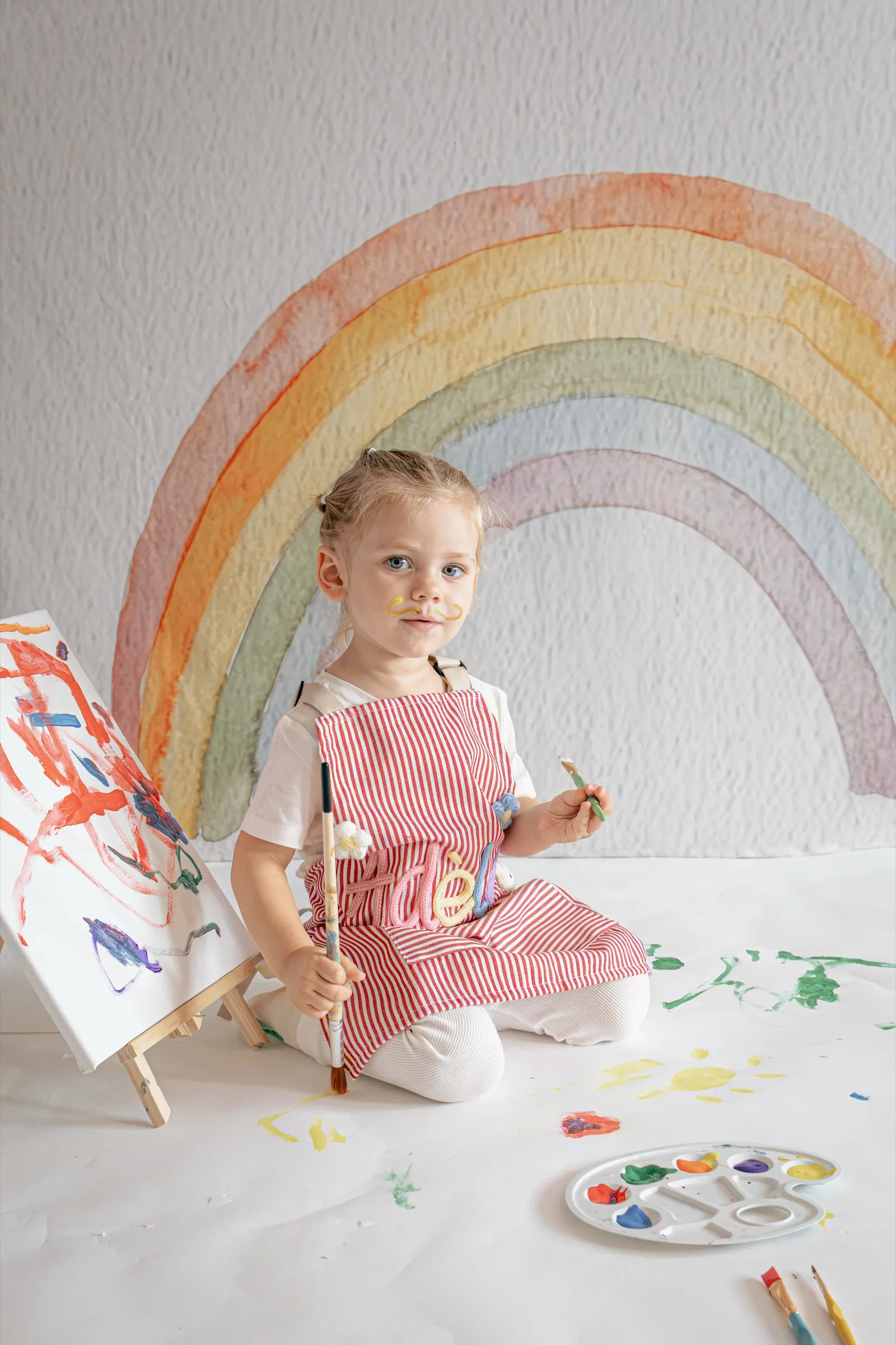 Petite fille en tablier rayé peignant sur un chevalet devant un décor arc-en-ciel, séance photo enfant en studio - Cindy Poulet photographe