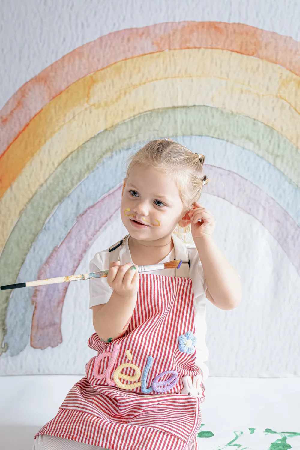 Petite fille au visage maquillé de peinture tenant son pinceau devant un décor arc-en-ciel, séance photo enfant - Cindy Poulet photographe