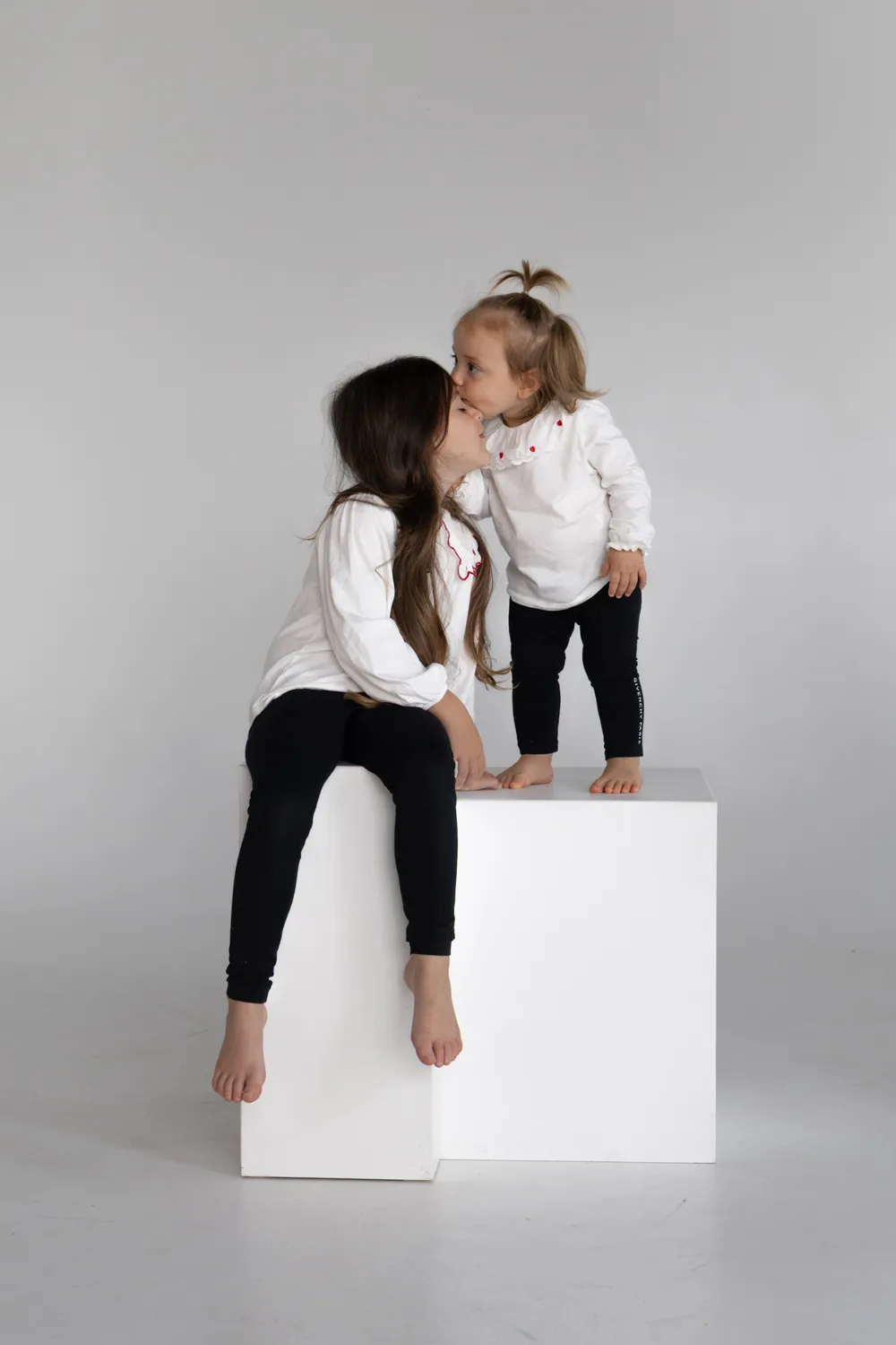 Deux sœurs en haut blanc et legging noir sur un cube blanc, la cadette embrasse le front de l'aînée, séance enfant en studio - Cindy Poulet photographe