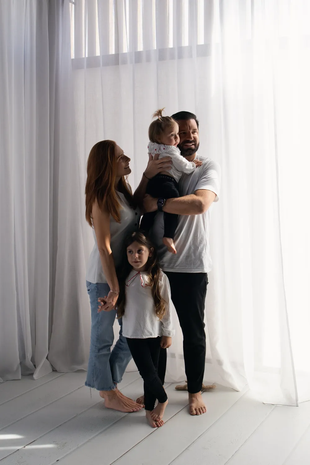 Parents et leurs deux filles debout pieds nus devant des voilages blancs, le papa porte la cadette, séance famille en studio - Cindy Poulet photographe