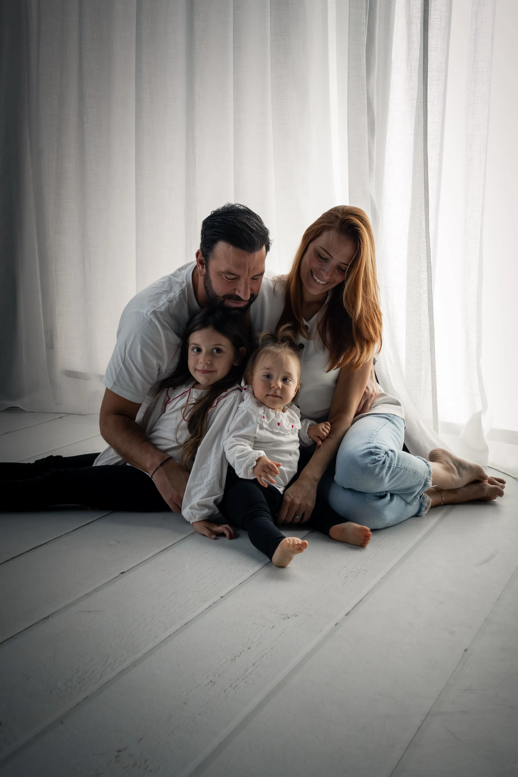 Parents et leurs deux filles blottis assis au sol devant des voilages blancs, séance famille en studio - Cindy Poulet photographe