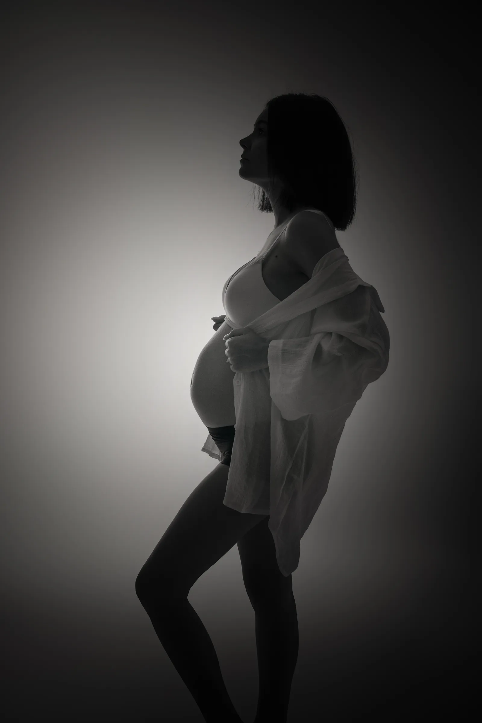 Femme enceinte de profil en contre-jour, chemise blanche ouverte sur son ventre rond, portrait maternité en noir et blanc - Cindy Poulet photographe