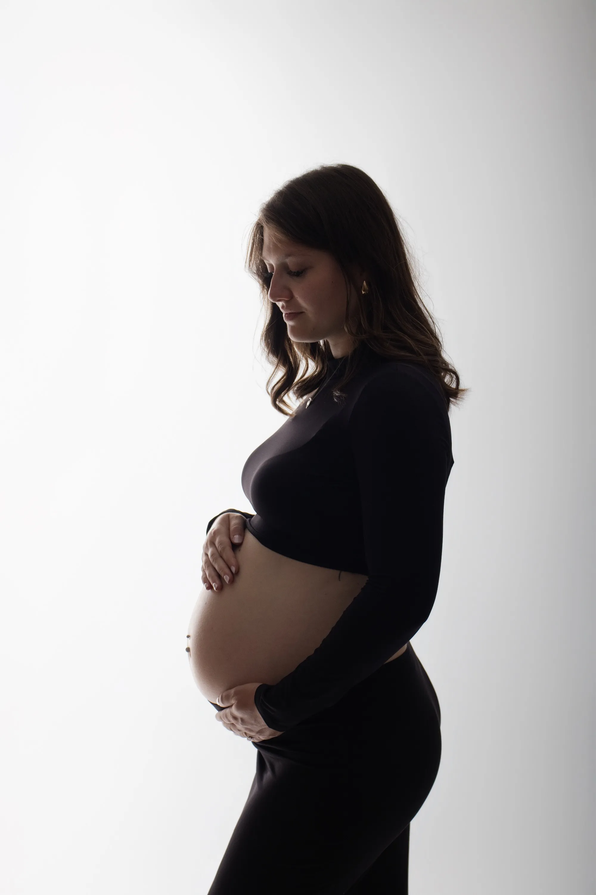 Femme enceinte de profil en tenue noire, mains sous son ventre rond, portrait maternité en studio - Cindy Poulet photographe