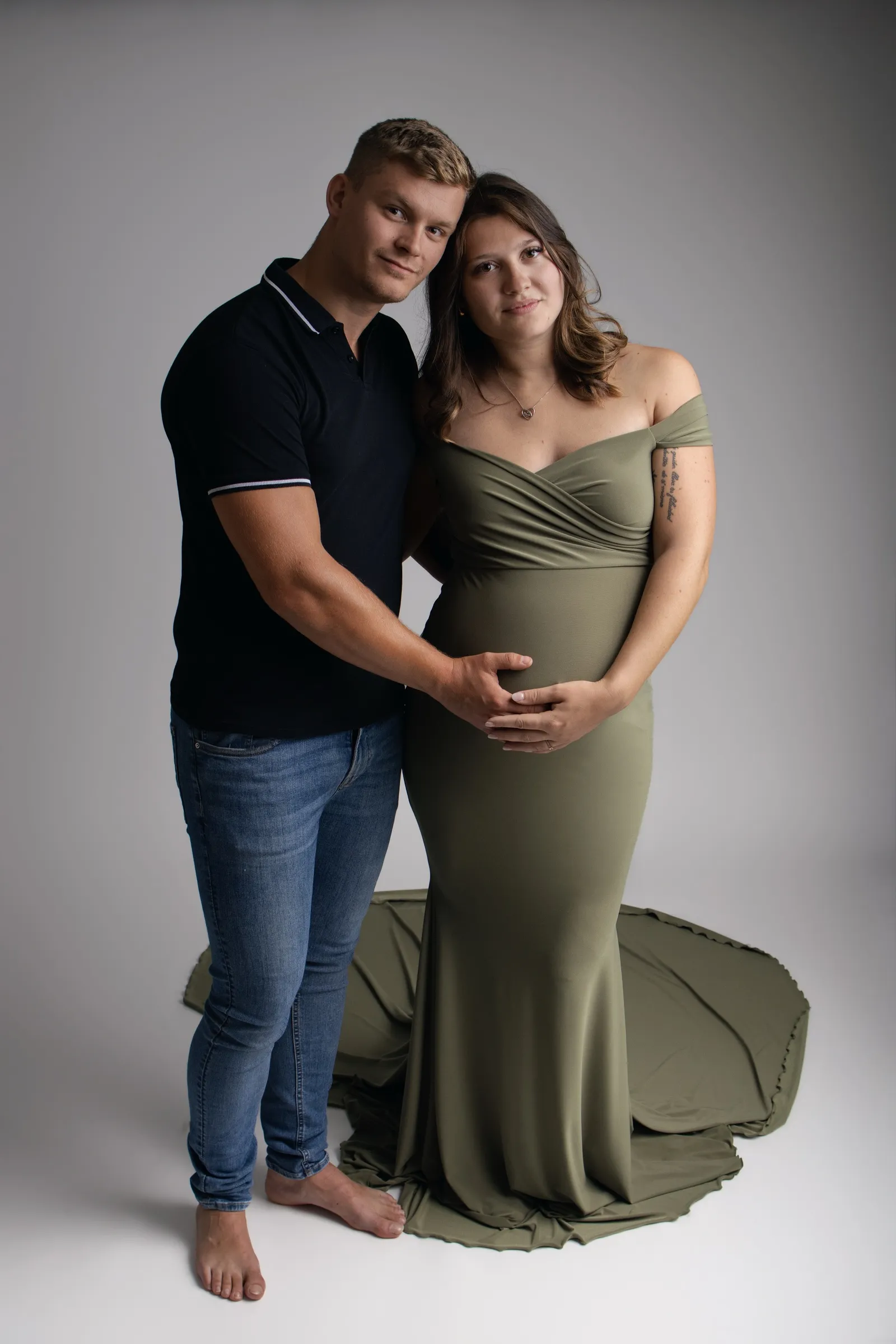 Couple posant en studio, future maman en robe longue vert sauge, mains réunies sur le ventre rond - Cindy Poulet photographe