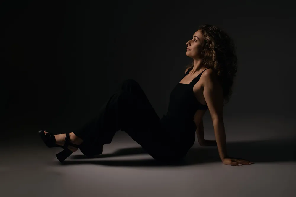 Femme en tenue noire assise au sol dans un studio sombre, portrait Élégance et Ombre - Cindy Poulet photographe