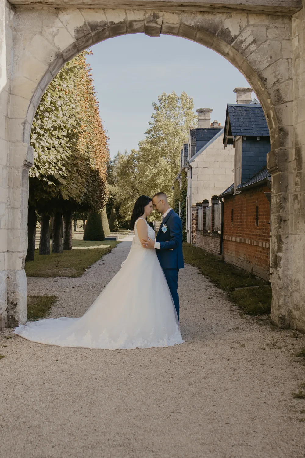 Les mariés enlacés front contre front sous une arche de pierre, au bout d'une allée de gravier - Cindy Poulet photographe