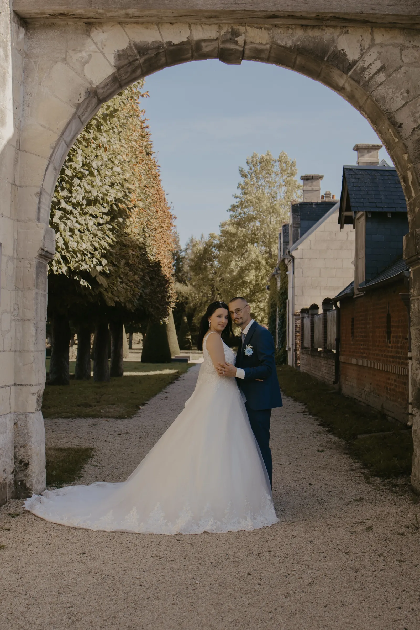 Couple de mariés enlacé sous une arche en pierre, regard vers l'objectif - Cindy Poulet photographe