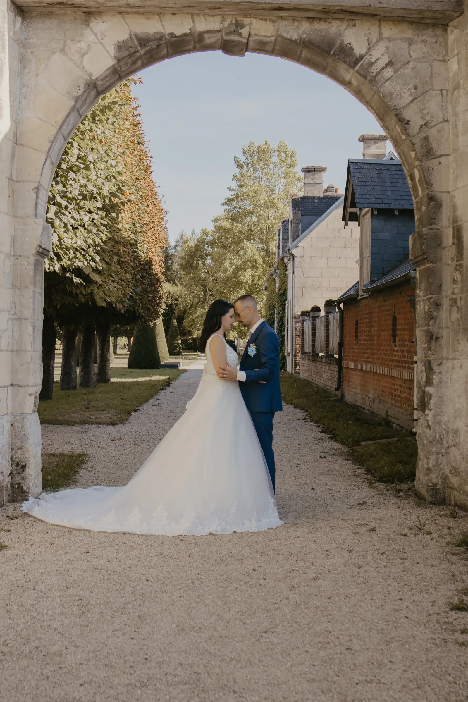 Mariés s'embrassant sous une arche en pierre, longue traîne déployée dans l'allée d'un château - Cindy Poulet photographe