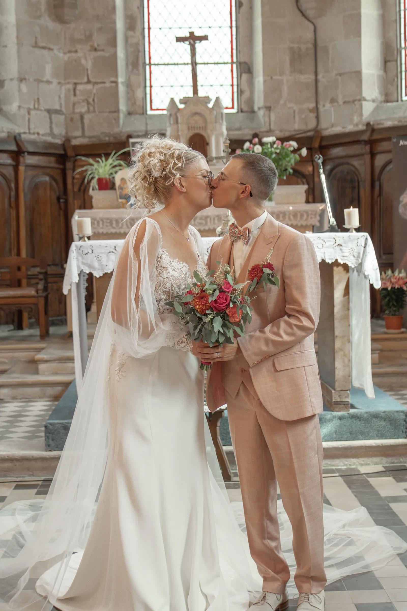 Baiser des mariés devant l'autel de l'église, mariée en robe de dentelle et marié en costume rose poudré - Cindy Poulet photographe