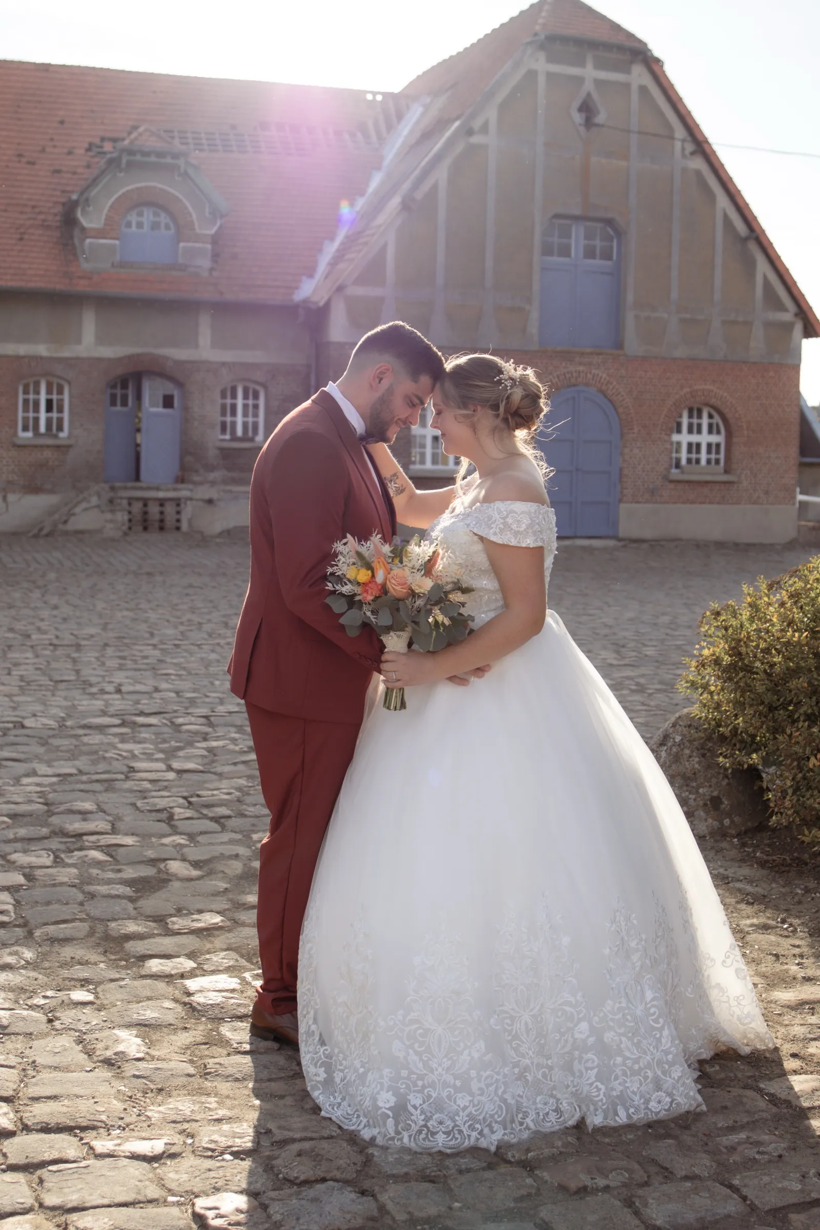 Mariés enlacés à contre-jour devant une longère en brique, marié en costume bordeaux et bouquet automnal - Cindy Poulet photographe