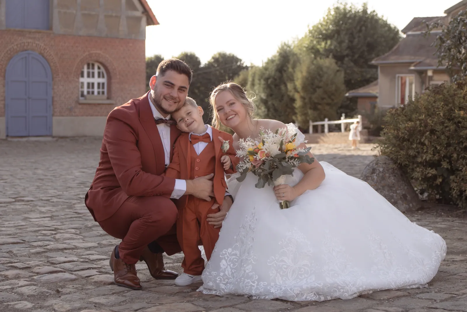 Les mariés accroupis avec leur petit garçon en costume terracotta dans une cour pavée - Cindy Poulet photographe