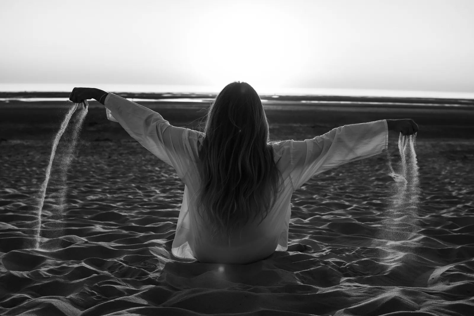 Femme de dos assise sur la plage, bras ouverts laissant couler le sable, portrait artistique en noir et blanc - Cindy Poulet photographe