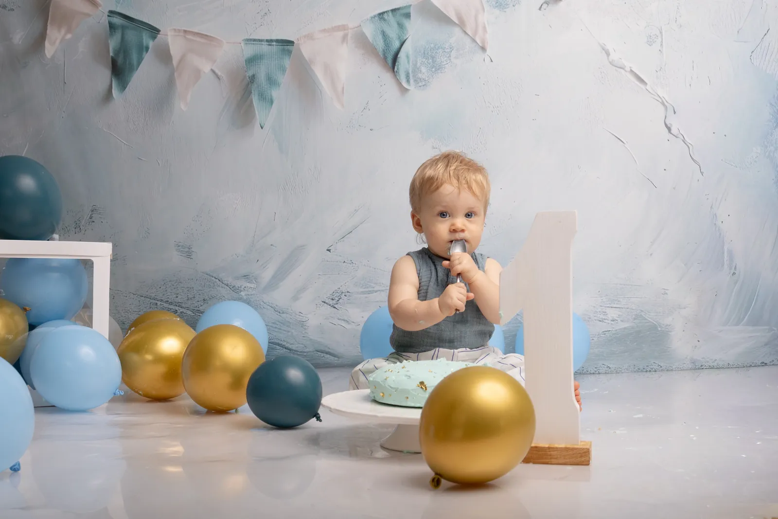 Bébé d'un an devant son gâteau de smash cake, ballons bleus et dorés et chiffre 1, séance anniversaire en studio - Cindy Poulet photographe