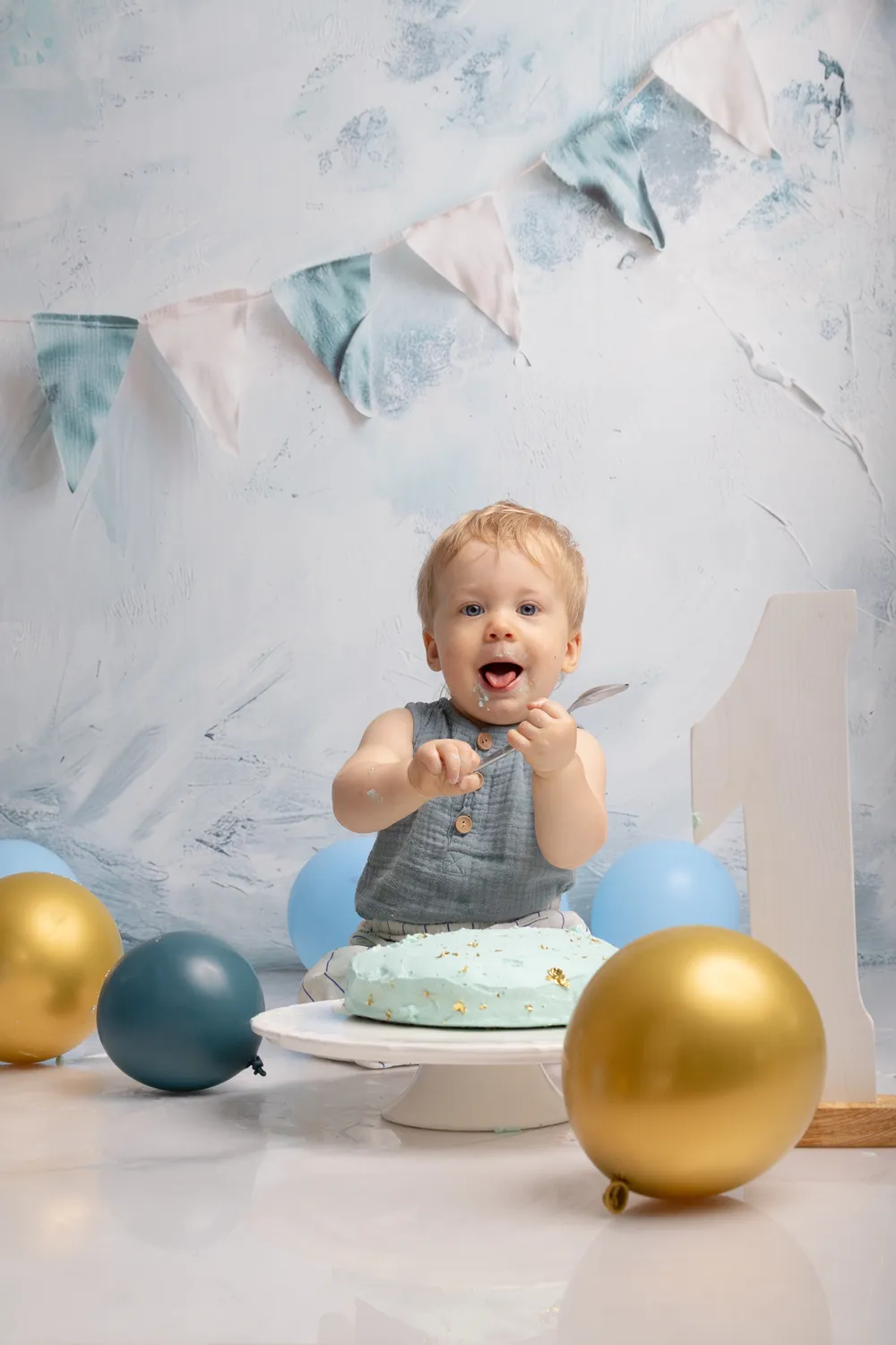 Bébé d'un an cuillère en main devant son gâteau vert d'eau, ballons dorés et bleus, chiffre 1 et fanions, smash cake en studio - Cindy Poulet photographe