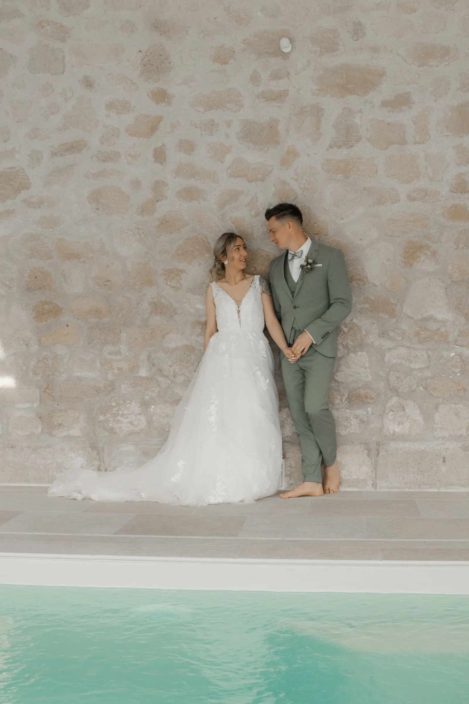Couple de mariés debout au bord d'une piscine devant un mur en pierre, photo de mariage - Cindy Poulet photographe