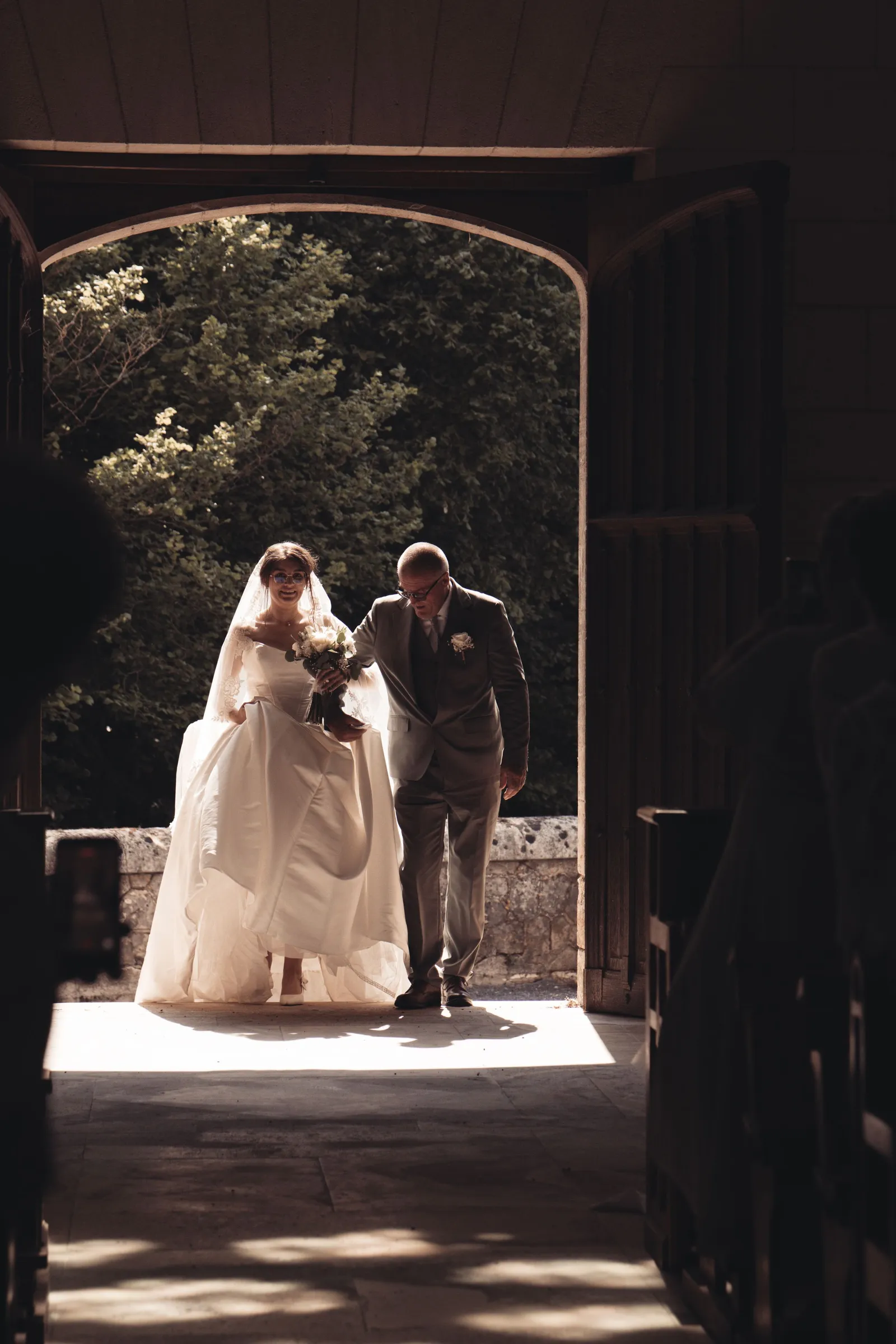 Entrée de la mariée au bras d'un proche sous un porche à contre-jour - Cindy Poulet photographe